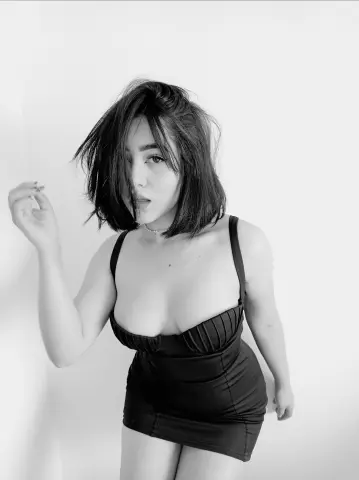AnnaKomatsuu poses in a black mini dress with a low-cut neckline against a plain white background.