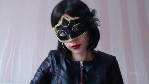 ViolenaSmith wears a black and gold masquerade mask and a black leather jacket against a light pink background.