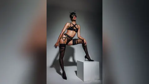 LucyMartin poses in black lingerie and sheer black stockings, standing with one leg raised on a white block against a grey background.