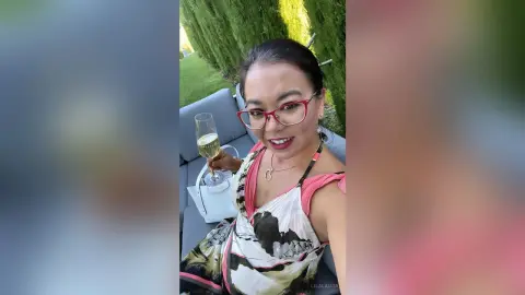 Smiling woman in a floral sundress holding a glass of white wine in a sunny outdoor garden setting.