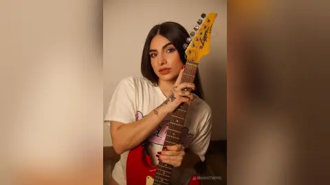 Young woman in a white graphic t-shirt holding an electric guitar in a minimalist studio setting.