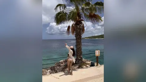 RossiNoleen stands on a stone walkway by the ocean, wearing a white bikini and a white open shirt, looking out at the sea near a palm tree.