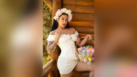 JuliahLewis wears a white lace corset and a floral headpiece while sitting against a rustic wooden log wall.