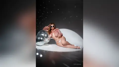 Glamorous woman in a pink faux fur coat and sunglasses posing with disco balls in a dreamy studio setting.