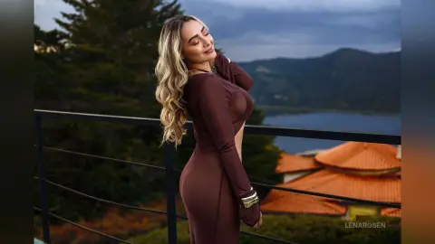Blonde woman in a brown long-sleeve dress posing on a balcony overlooking a scenic mountain lake at dusk.