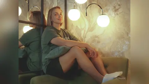 Smiling young woman in a green t-shirt relaxing on a velvet sofa in a warm, dimly lit room.