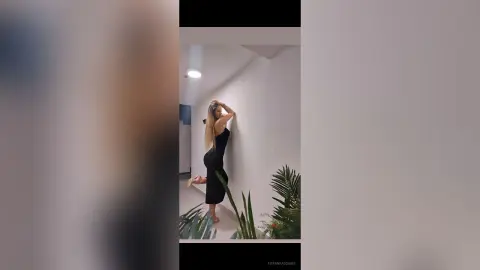 TiffanyAddams poses against a white wall in a black sleeveless dress, running her hands through her long blonde hair.