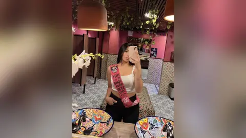 Young woman in a white tank top and pink sash taking a selfie in a cozy, plant-filled restaurant setting.