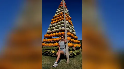 Smiling woman in a grey t-shirt and denim shorts sitting by a massive stack of pumpkins under a clear blue sky.