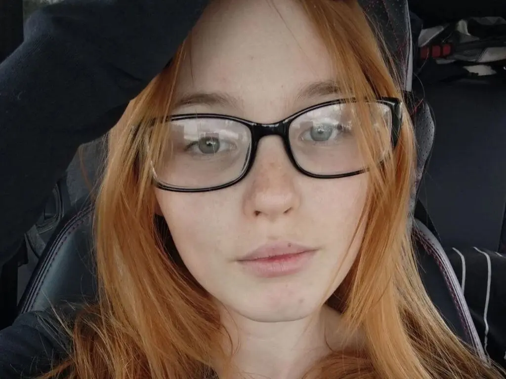 VetaFerr is a redhead with glasses looking directly into the camera while sitting in a car seat.
