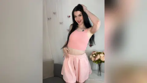 Smiling young woman in a pink ribbed tank top and shorts posing in a bright, minimalist room.