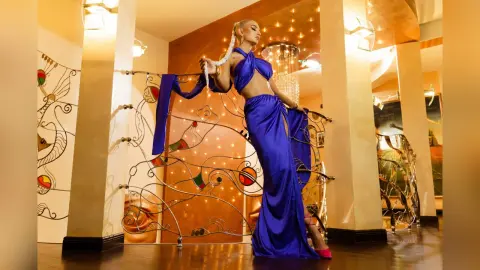 Anastasia poses in a long blue silk dress while leaning against a decorative metal railing in a warm, lit room.