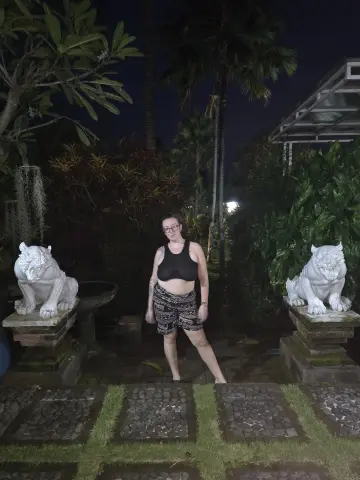 Woman in black crop top and patterned shorts standing in a tropical garden at night near stone lion statues.