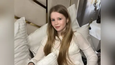 Blonde woman in a white long-sleeve shirt relaxing on a white pillow in a bright, cozy bedroom setting.