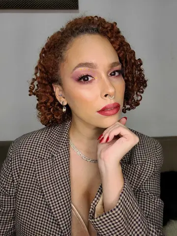 dominamoon — Curly-haired woman in a brown houndstooth blazer and red lipstick posing with a confident expression.