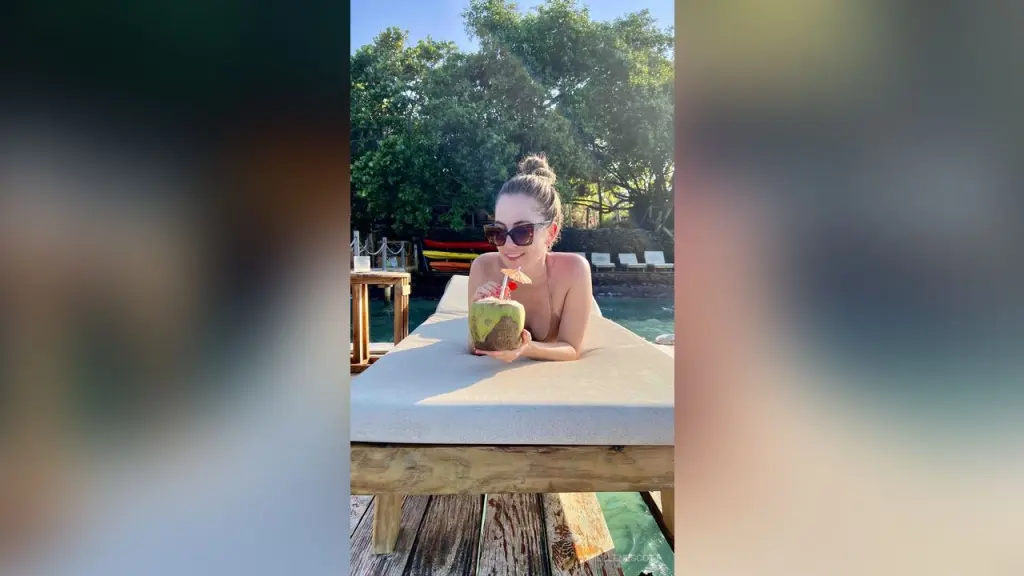 SeleneScott — Smiling woman in sunglasses enjoying a fresh coconut drink on a sunny tropical resort lounge chair.