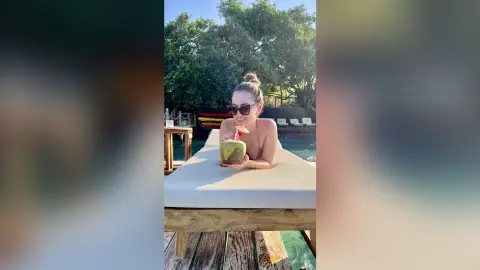 Smiling woman in sunglasses enjoying a fresh coconut drink on a sunny tropical resort lounge chair.