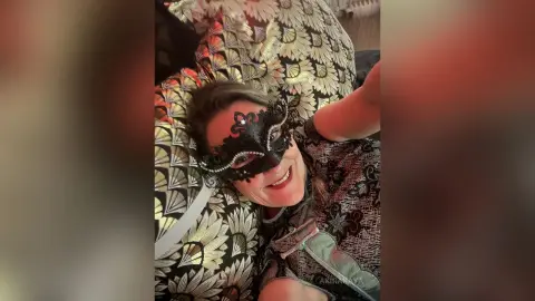 Smiling woman in a black masquerade mask and patterned outfit relaxing on a gold and black patterned cushion.