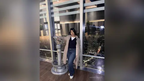 Young woman in a beige coat and jeans standing by a window overlooking a glowing city skyline at night.