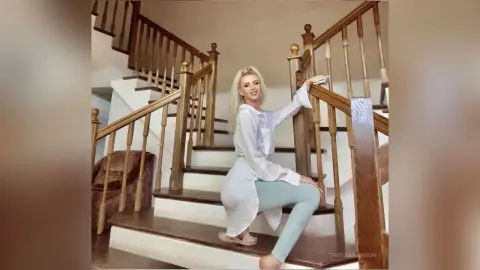 Blonde woman in a white button-down shirt and light blue jeans relaxing on a wooden staircase in a bright home.