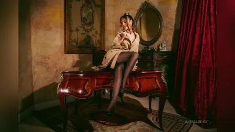 Elegant woman in a beige silk robe sitting on a vintage red desk in a moody, classic luxury room.