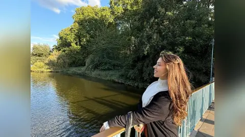 Smiling woman in a white turtleneck and black sweater enjoying a sunny day by a peaceful lake.