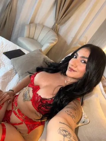 Smiling woman with long dark hair wearing red lace lingerie while relaxing on a white bed in a bright room.