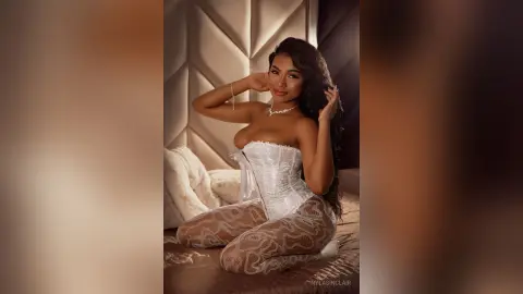 Smiling woman in white lace corset and sheer patterned leggings relaxing on a cozy bed in a warm bedroom setting.