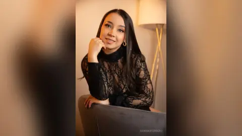 Smiling woman in a black lace long-sleeve top sitting in a warm, cozy indoor setting.