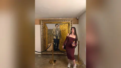 Stylish woman in a burgundy midi dress standing in front of a large framed portrait in a modern art gallery.