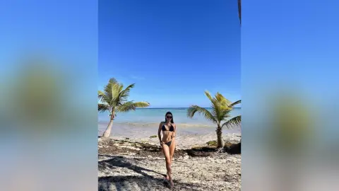 Woman in a black bikini standing on a sunny tropical beach with palm trees under a clear blue sky.