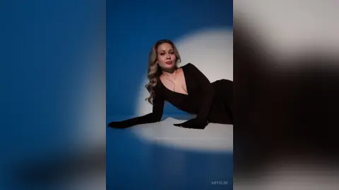 Elegant blonde woman in a chic black long-sleeve dress posing against a moody blue studio background.