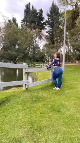 Woman in floral blouse and blue jeans leaning on a wooden fence by a peaceful lake in a lush green park.