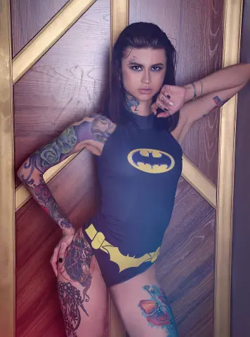 Tattooed woman in a black Batman bodysuit posing against a wooden door in a moody, cinematic setting.