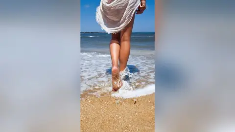MarisaGrace stands on a sandy beach with the ocean waves washing over her bare feet.