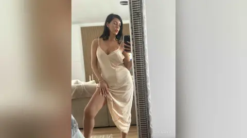 Young woman in a champagne silk slip dress posing for a mirror selfie in a bright modern bedroom.