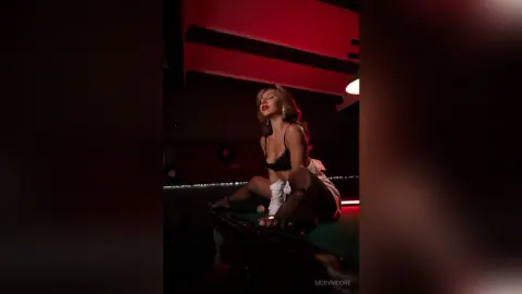 Glamorous woman in black lingerie and white robe sitting in a moody, red-lit lounge setting.