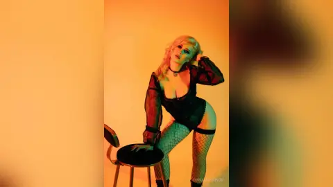 Blonde woman in black lace bodysuit and fishnet tights posing in warm orange studio lighting.