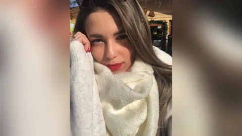 MontseFerrera poses with her hand near her face, wearing a thick white knit scarf or sweater against a soft outdoor background.