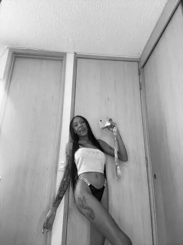 Smiling tattooed woman in a white crop top and black underwear taking a selfie with an instant camera indoors.