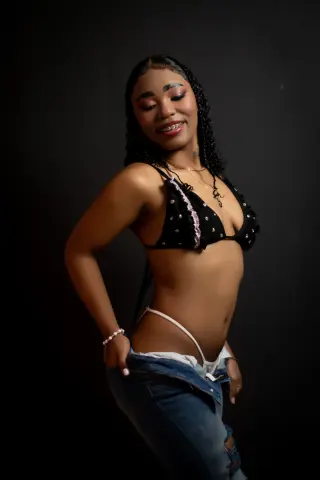 Smiling young woman in a black bikini top and denim jeans posing against a dark studio background.