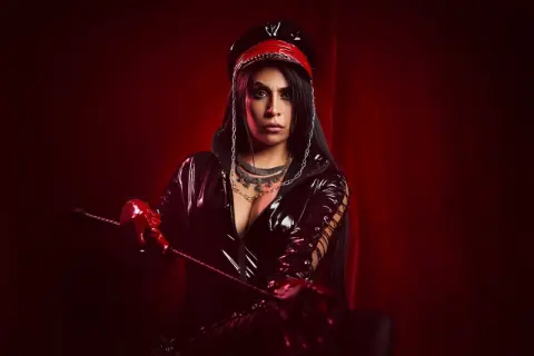 Moody portrait of a woman in a black latex outfit and red cap holding a whip against a dark red background.