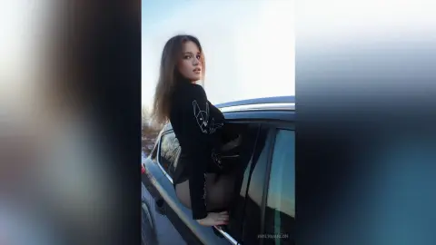 Young woman in a black long-sleeve top sitting in a car window during a bright outdoor sunset.