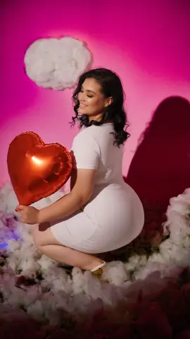 Smiling woman in a white dress holding a red heart balloon amidst dreamy pink clouds.
