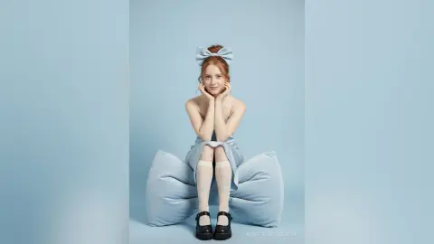 Redheaded woman in a light blue dress and bow sitting on a beanbag against a blue background with a serene mood.