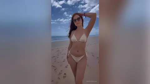 Young woman in a cream bikini and sunglasses posing on a sunny beach under a blue sky.