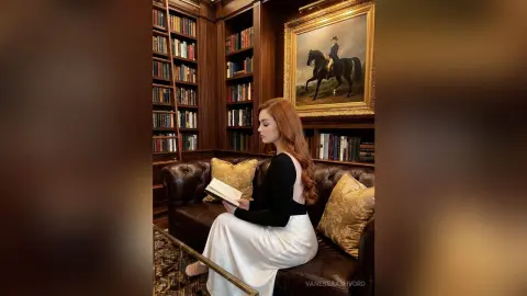 Redheaded woman in a black backless top and white skirt reading a book on a leather sofa in a classic library.
