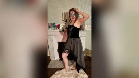 Woman in black lace lingerie and mask posing with a red rose in a cozy living room setting.