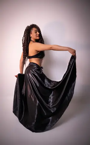 Elegant woman in a black satin crop top and flowing maxi skirt posing against a minimalist studio background.