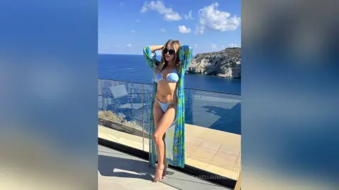 Stylish woman in a blue bikini and sheer floral cover-up posing on a sunny seaside balcony overlooking the ocean.